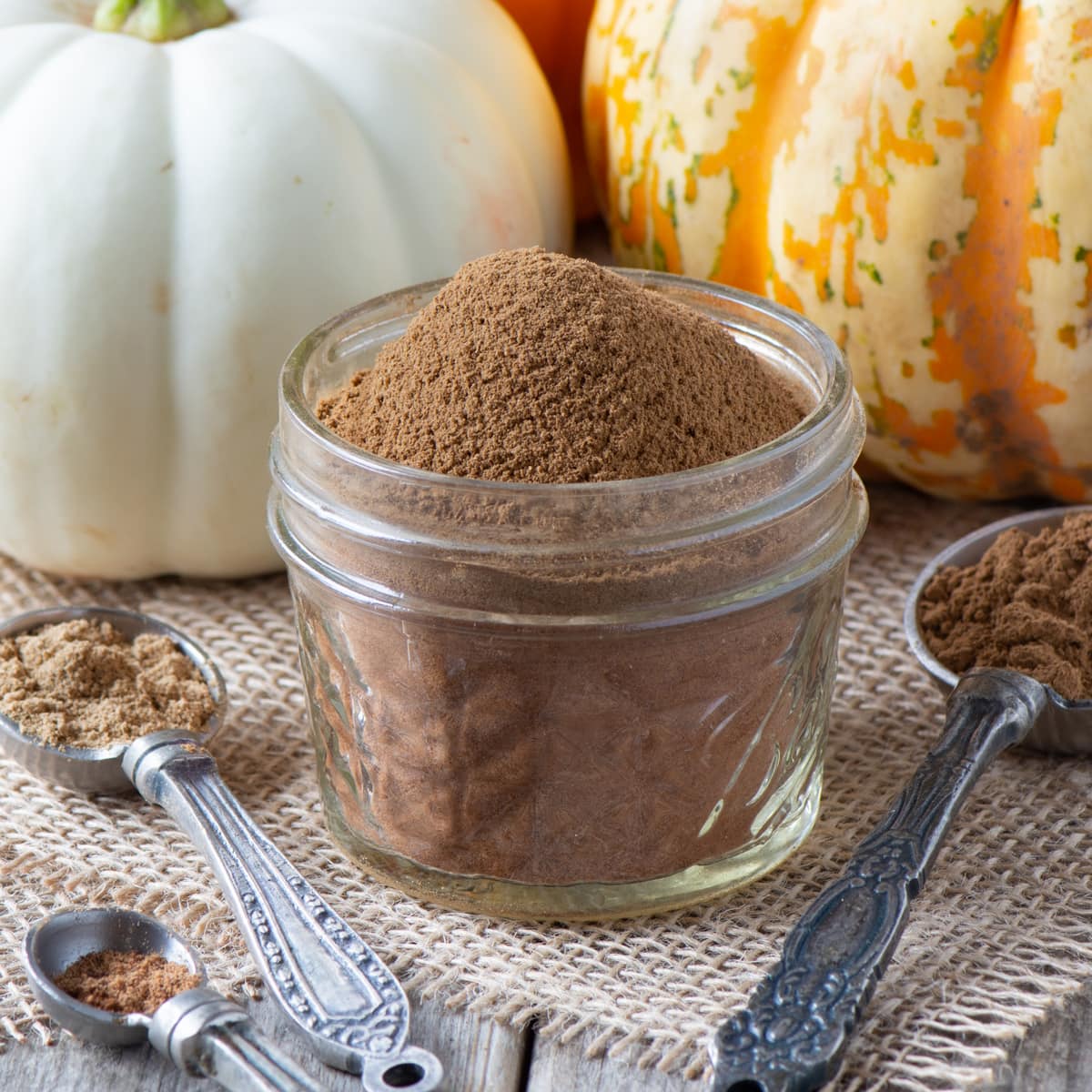 View of a small jar of pumpkin spice with the contents protruding from the top with spoons filled with the ingredients in the front and pumpkins in the background. 