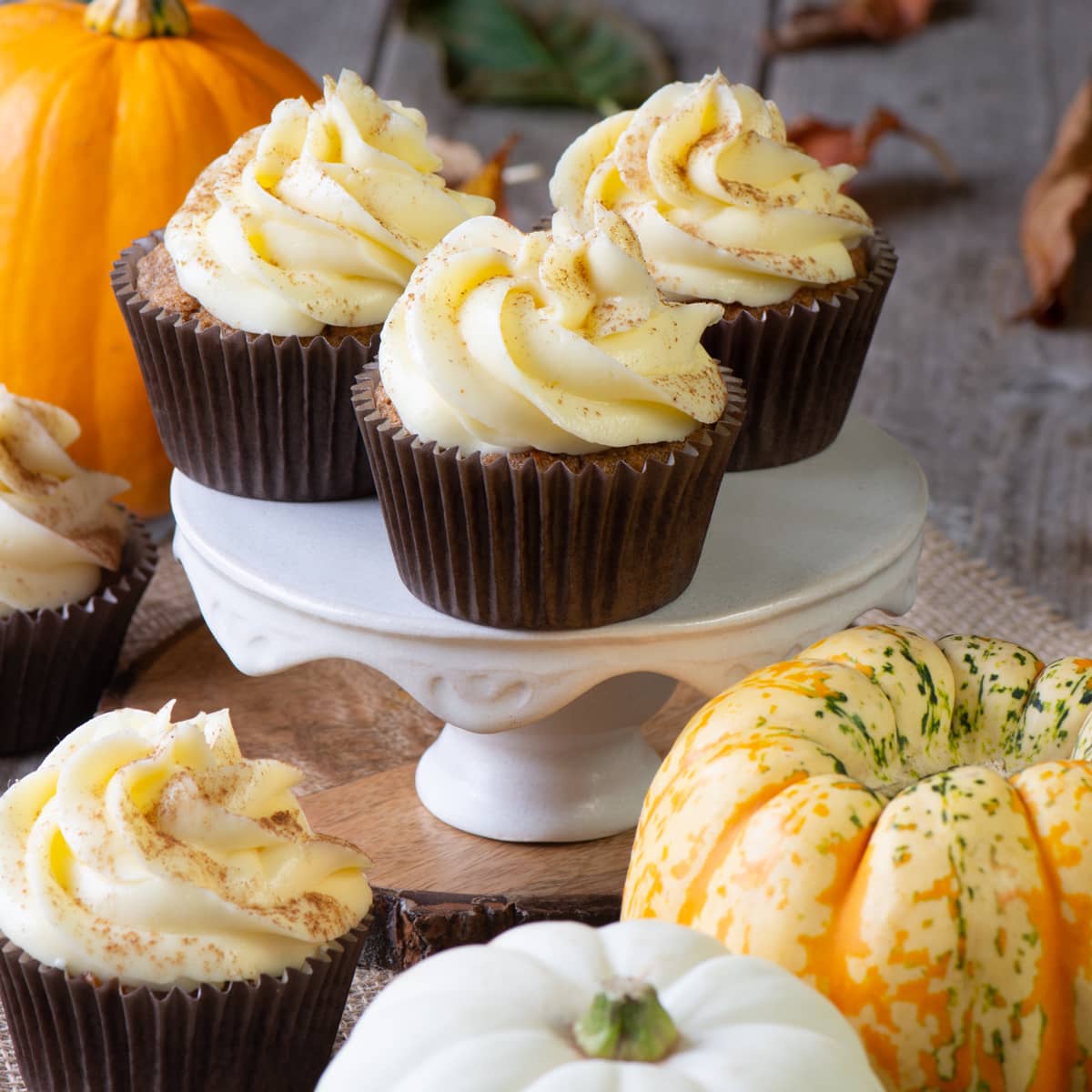 Three frosted pumpkin cupcakes shown on a white cake stand surrounded by small pumpkins.  