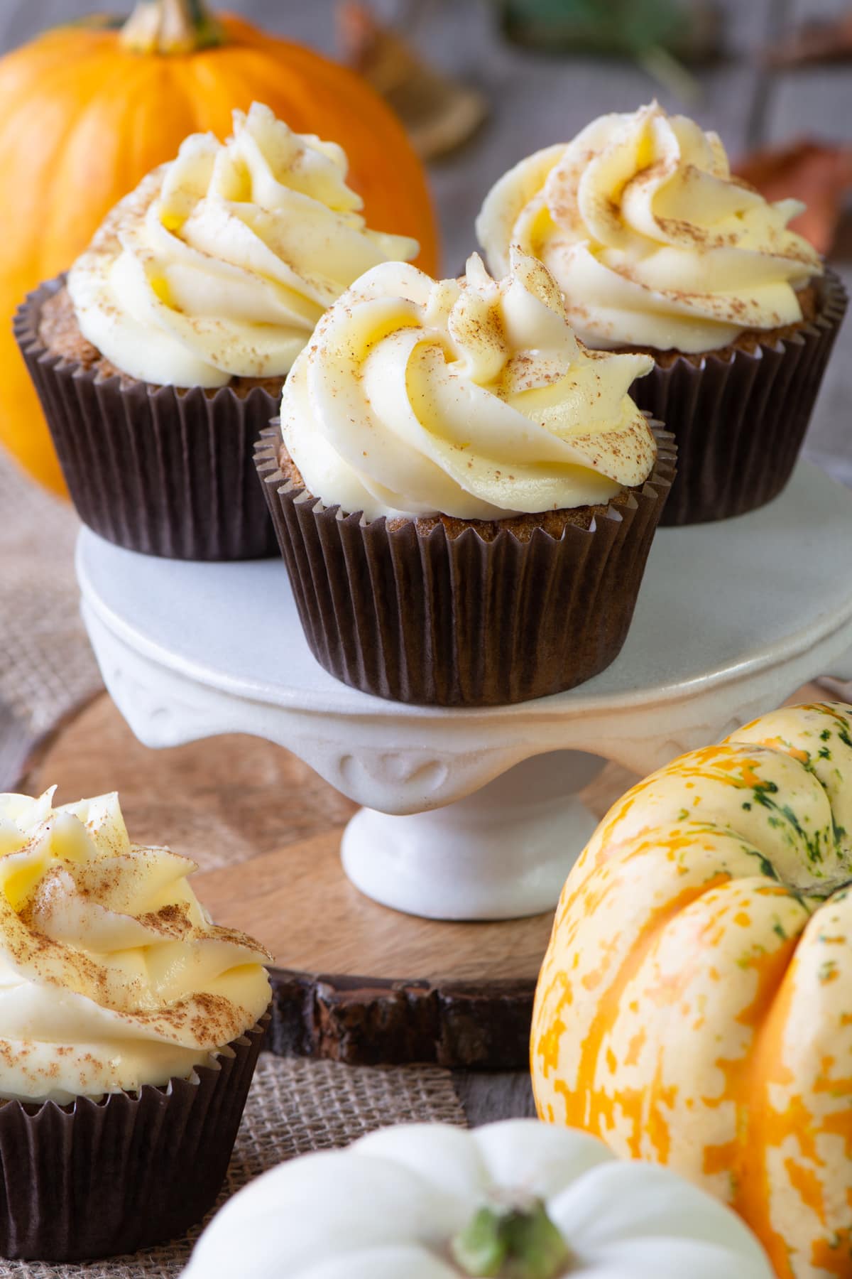 3 cupcakes shown on a cake stand with another cupcake shown in front with several small pumpkins in view. 